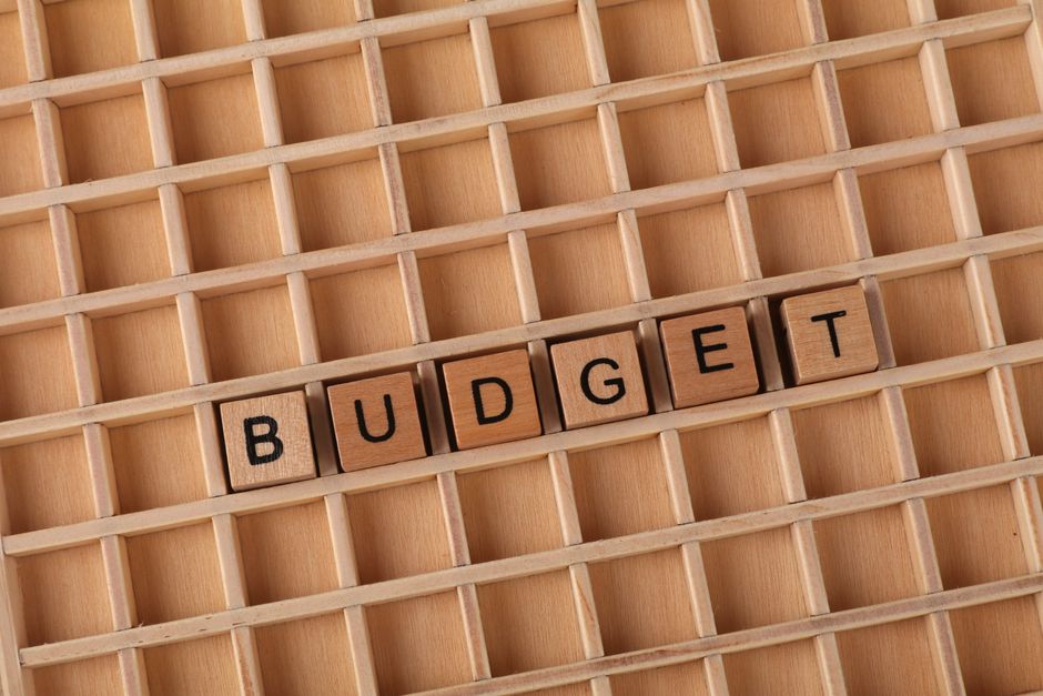 Close-up of a wooden grid or organizing tray with multiple small compartments, some of which are empty, while a row of wooden letter tiles spelling out the word 'BUDGET' is placed within one row. The